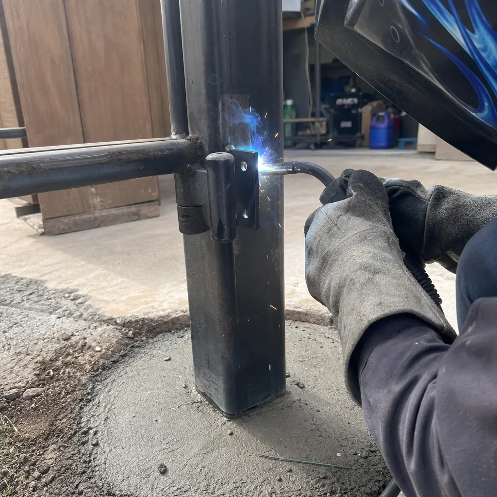 Welder installing a steel gate hinge onto a metal support post